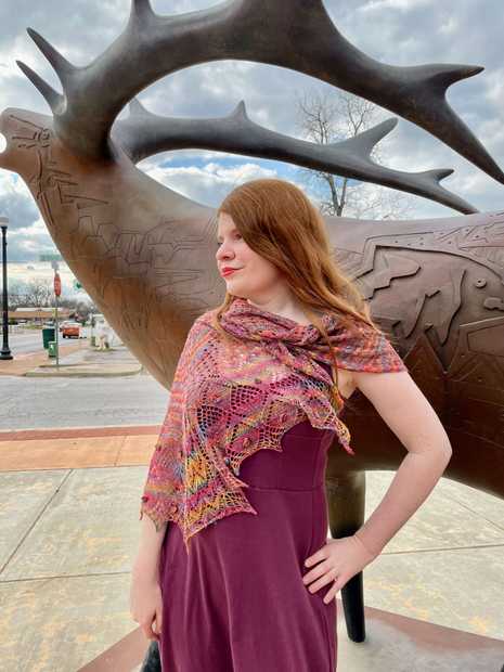 woman in brightly colored asymmetric lace shawl, standing in front of metal deer sculpture