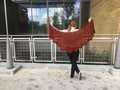 woman holding dark red Feuille-morte shawl suspended between her hands to demonstrate flat shape, and standing in front of railing at Austin Public Library